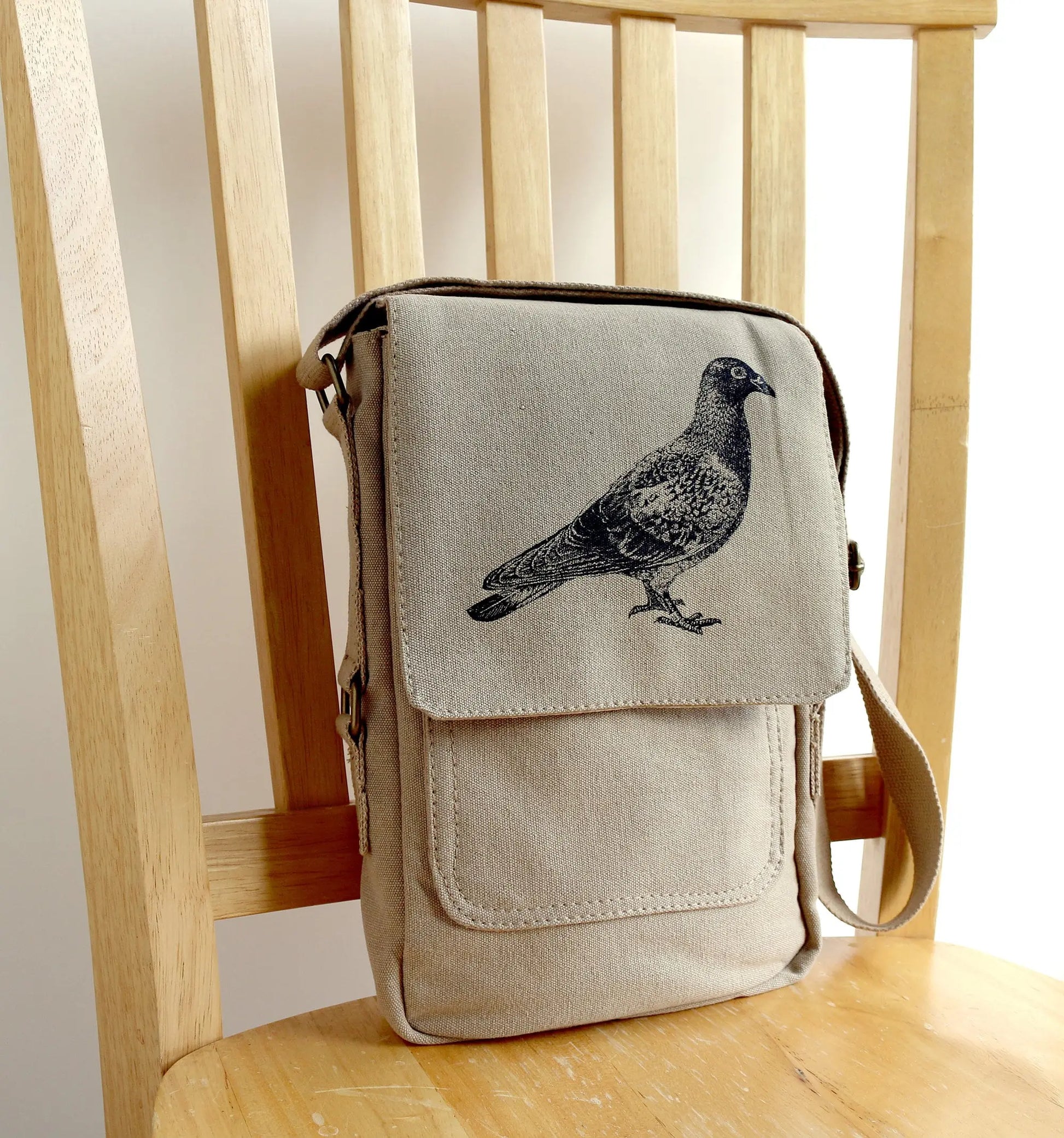 Beige canvas crossbody bag with black pigeon print, displayed on a wooden chair.