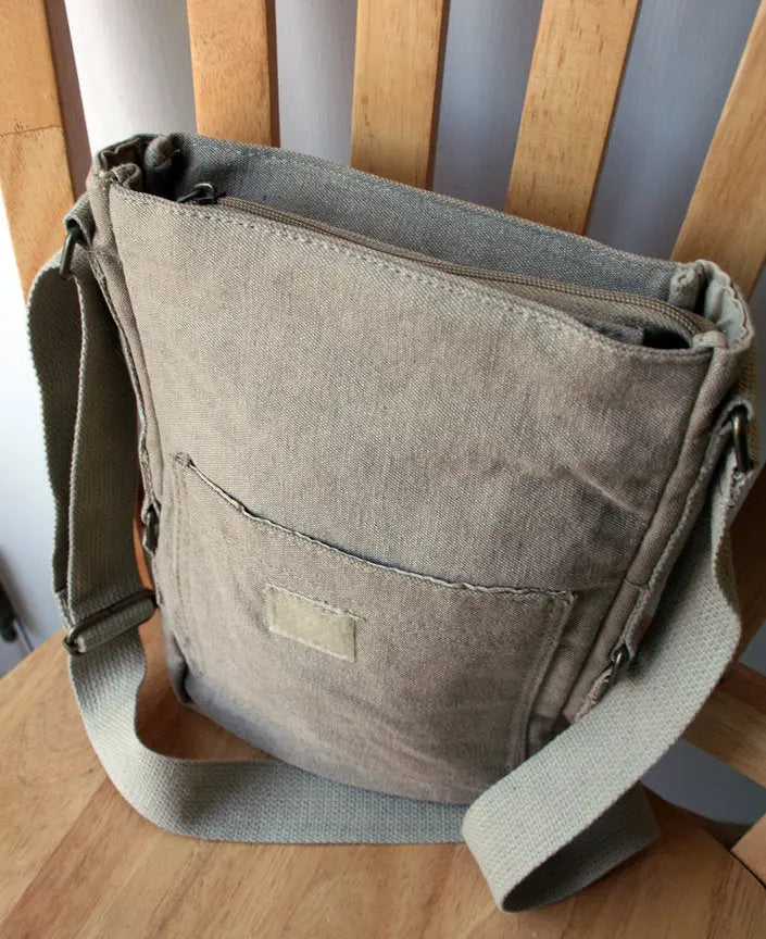 Beige canvas crossbody bag with front pocket and adjustable strap on a wooden chair.