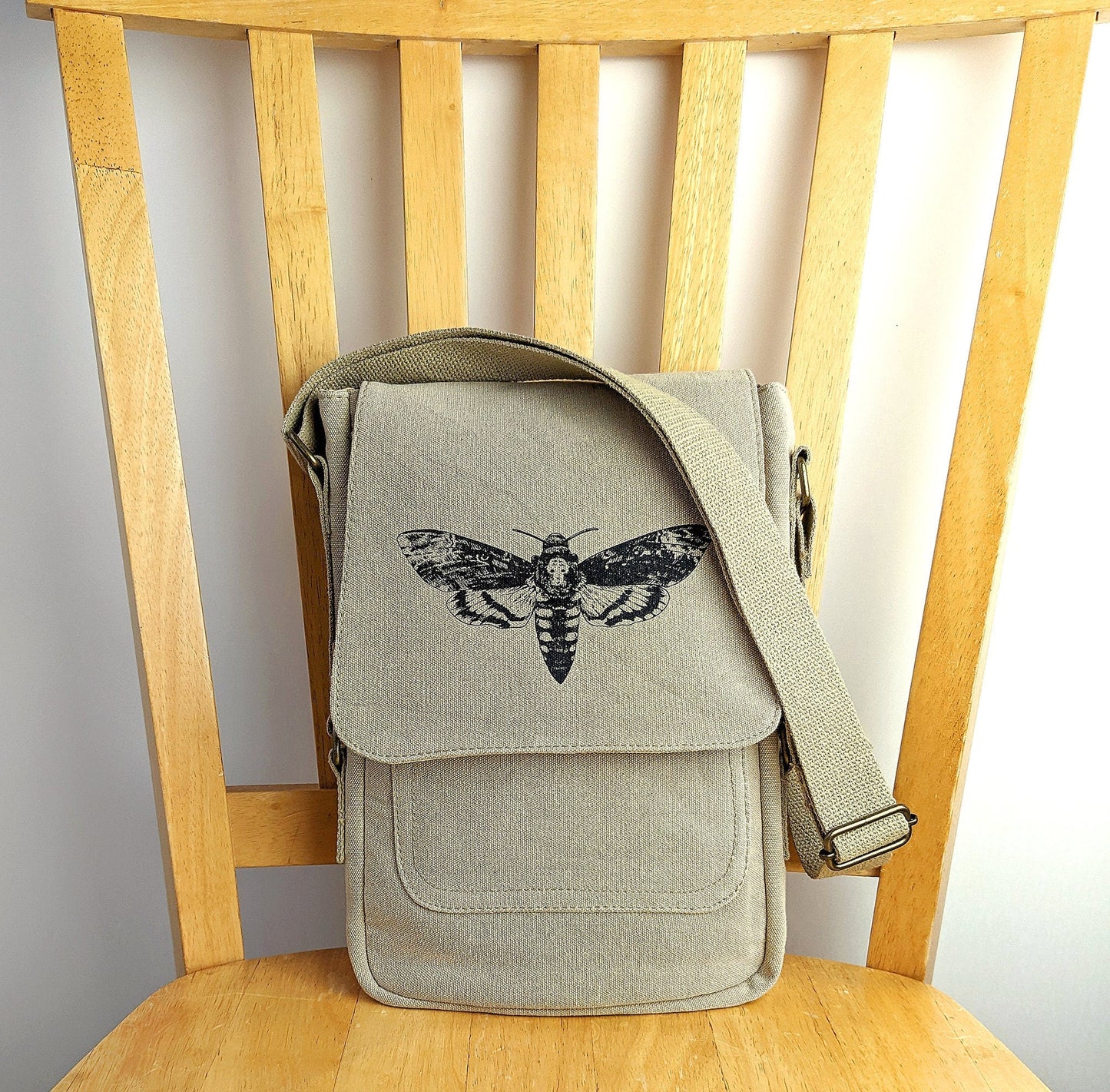 Beige canvas shoulder bag with black moth screen print, displayed on a wooden chair.