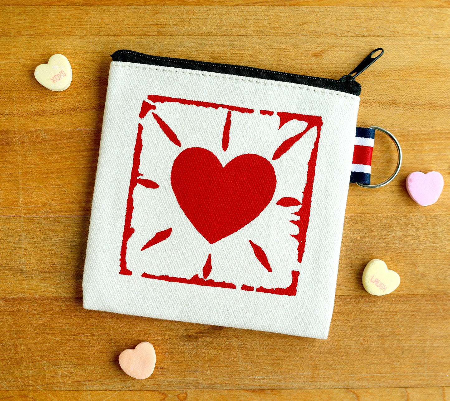 Hand-printed canvas zipper pouch with red heart design, surrounded by candy hearts on wood.