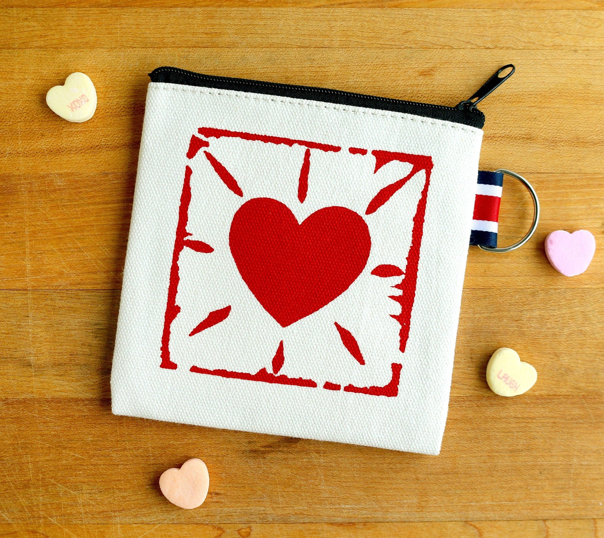 Hand-printed canvas zipper pouch with red heart design, surrounded by candy hearts on wood.