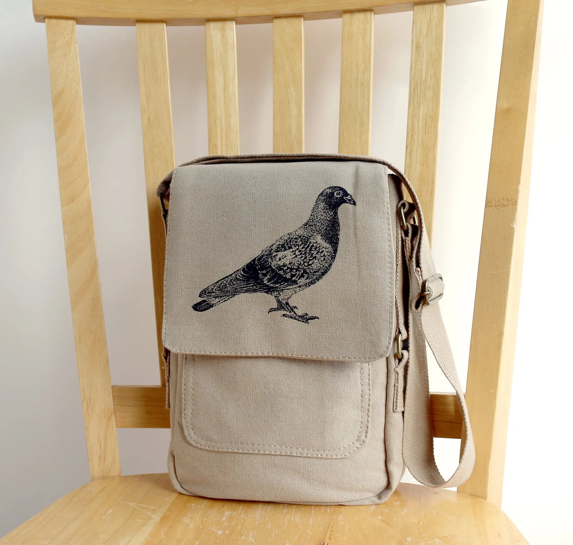 Beige canvas crossbody bag with black pigeon print, displayed on a wooden chair