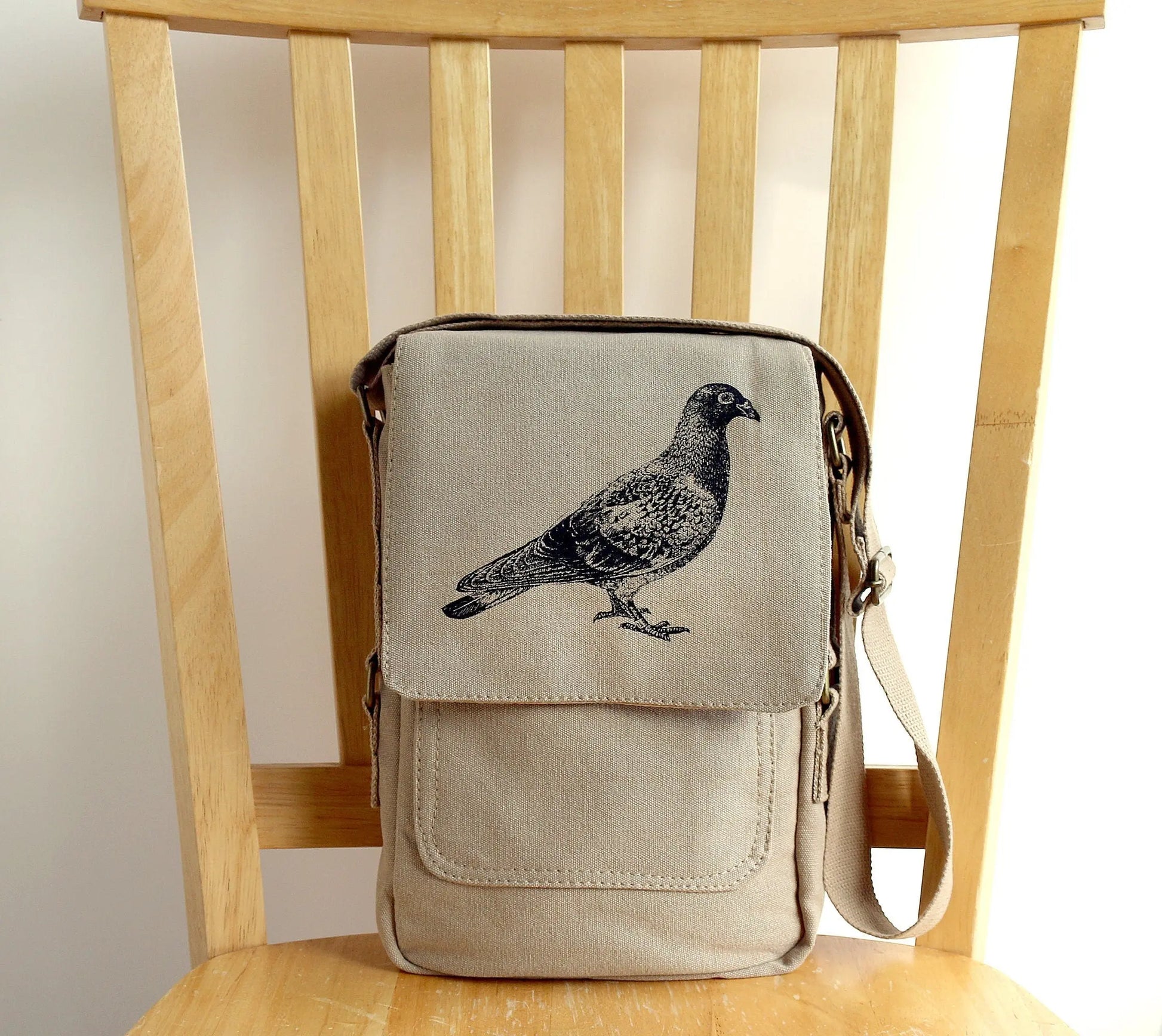 Beige canvas crossbody bag with black pigeon print on flap, displayed on wooden chair.