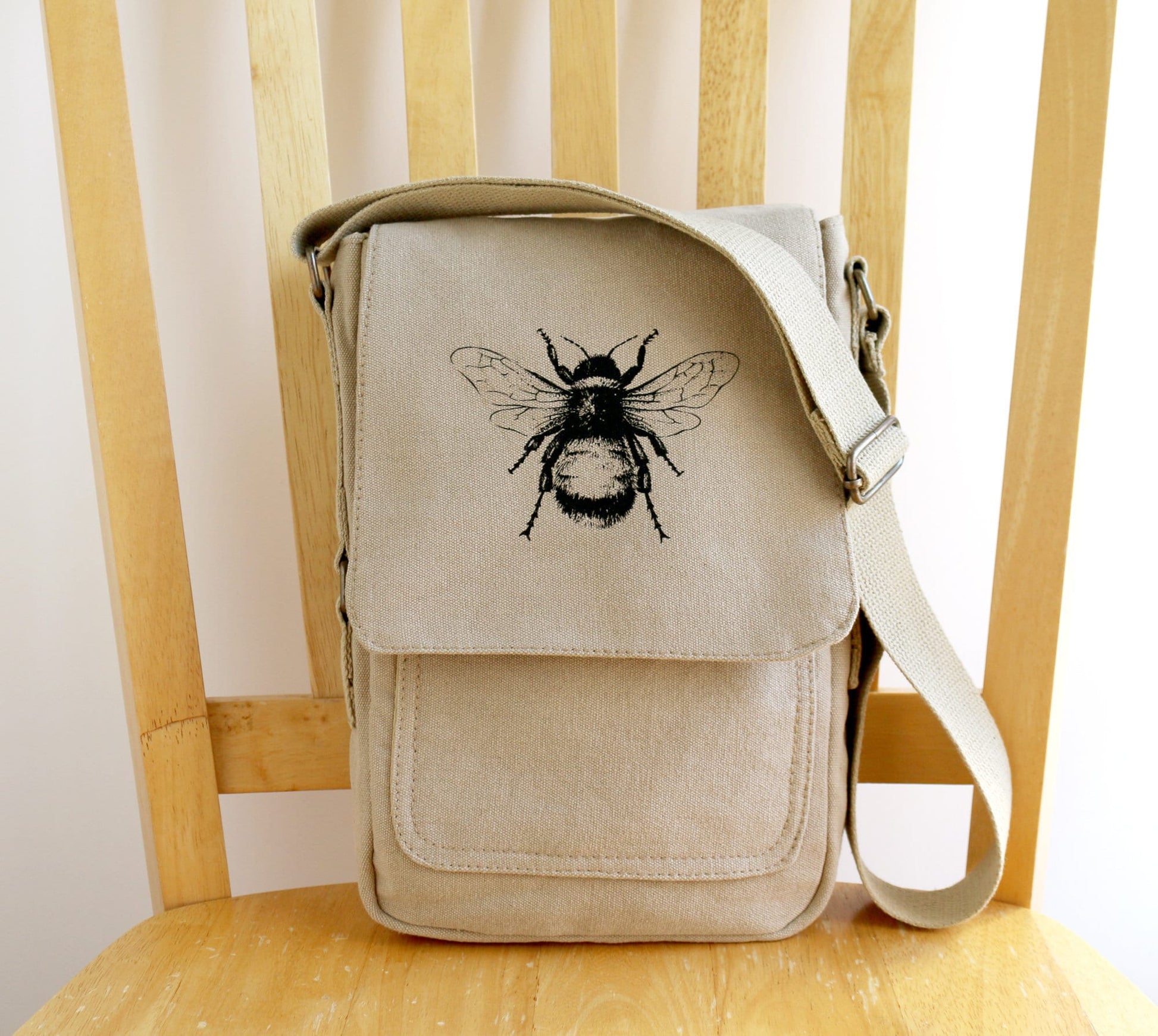 Beige canvas crossbody bag with black bee illustration, hand printed, on wooden chair