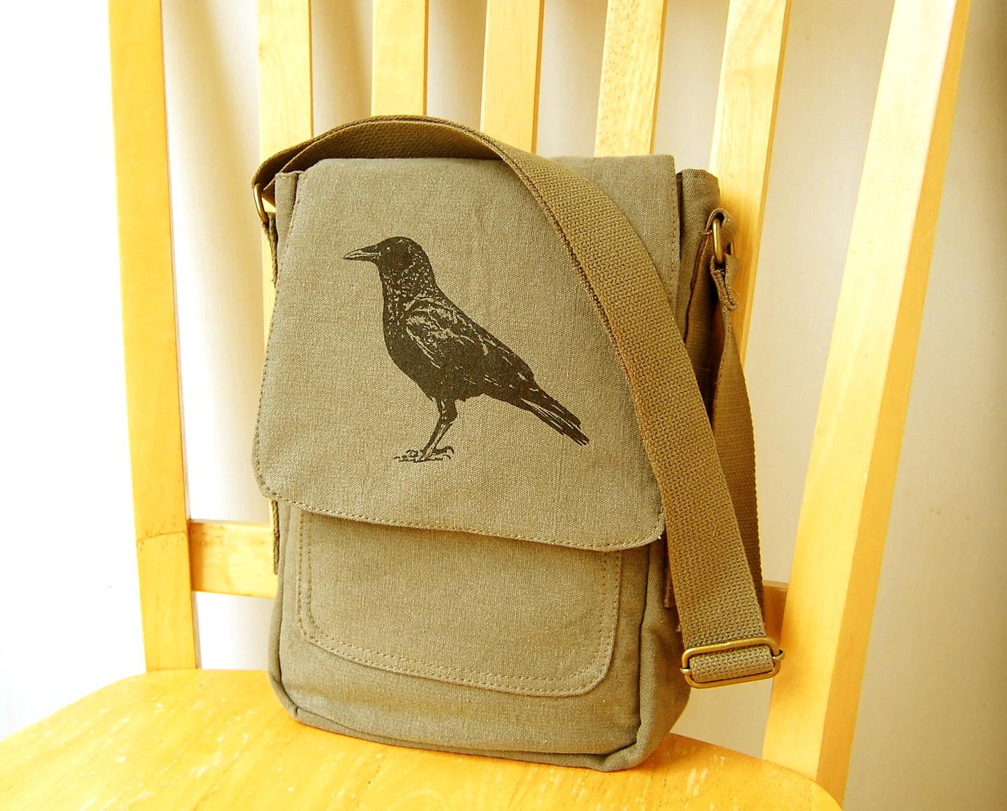 Olive green canvas crossbody bag with black crow print, displayed on a wooden chair.