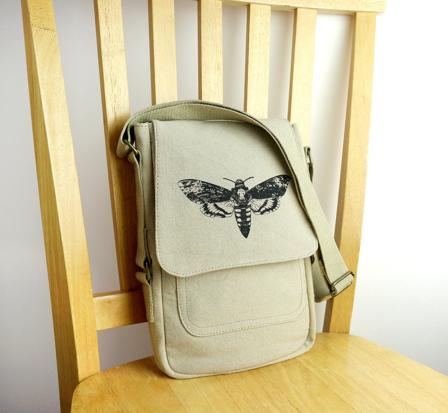 Beige canvas crossbody bag with black moth print, resting on a wooden chair