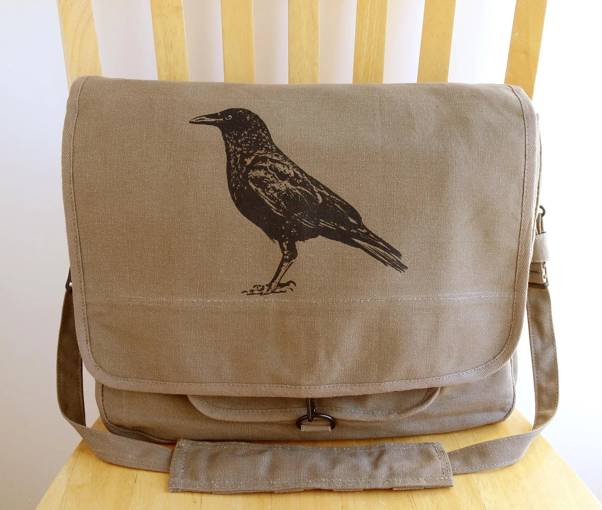 Brown canvas messenger bag with black screen-printed crow design on front flap, on chair